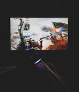 A dimly lit room with a television screen displaying a dynamic, action-packed video game interface featuring a historical battle scene. The foreground shows the silhouette of a gaming console controller held by a person.