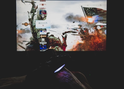 A dimly lit room with a television screen displaying a dynamic, action-packed video game interface featuring a historical battle scene. The foreground shows the silhouette of a gaming console controller held by a person.