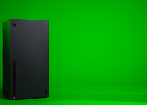 A black, rectangular gaming console stands upright against a vibrant green background. The console has a simple, sleek design with minimalistic features including a disc slot and a few buttons.