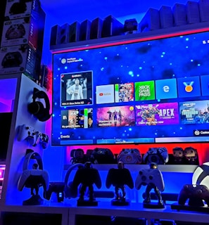 A modern gaming setup featuring a large wall-mounted screen displaying a gaming console interface. The room is decorated with gaming-themed items such as controllers and collectibles. The ambient lighting includes shades of blue and red, creating a vibrant atmosphere.