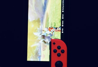 A gaming console with a screen displaying a video game scene, accompanied by two controllers, one colored red and the other blue. The devices are set against a dark backdrop, emphasizing the vibrant colors of the controllers.