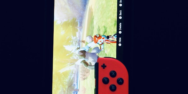 A gaming console with a screen displaying a video game scene, accompanied by two controllers, one colored red and the other blue. The devices are set against a dark backdrop, emphasizing the vibrant colors of the controllers.