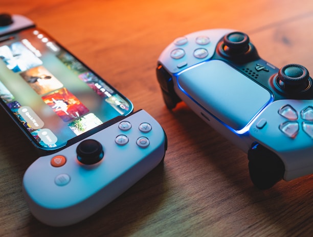 A gaming scenario featuring a smartphone with a game interface next to two controllers on a wooden surface. The smartphone displays vibrant images and icons, while the controllers feature a sleek, modern design with buttons and joysticks prominently visible. Warm lighting enhances the scene.