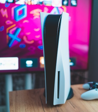 A gaming console is placed on a wooden table with a game controller nearby. In the background, a television screen displays colorful graphics, including geometric shapes and vibrant colors.