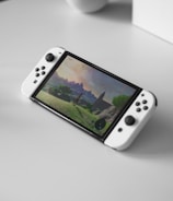 A portable gaming console with a screen showing a video game landscape, resting on a light-colored surface. The console has white controllers attached to either side of the screen. Nearby, there seems to be a white docking station or accessory.
