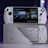 A handheld gaming console with a screen displaying a game library menu is placed on top of its packaging. The console features various buttons, a joystick, and a circular light around the left joystick. The background is blurred with purple, yellow, and blue tones.