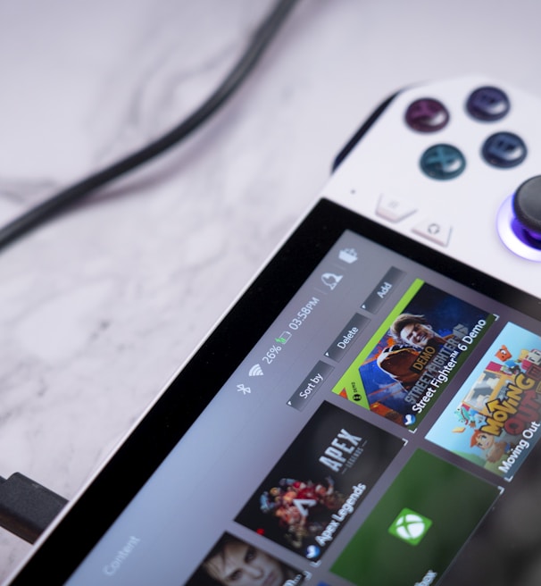 A handheld gaming console with a white body and colorful buttons features a screen displaying various game icons. The device is connected to a power source via a black cable and rests on a marble surface. The interface shows games like Apex and Street Fighter.