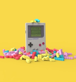 A vintage handheld gaming console surrounded by colorful, block-like pieces reminiscent of a classic puzzle game. The console has a small screen displaying a game in progress, with two red buttons and a directional pad. The vibrant pieces are scattered around it, creating a playful and nostalgic atmosphere.